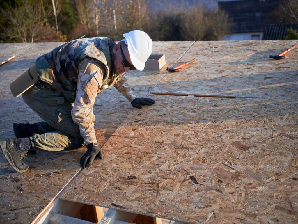 Roofer in a white hard hat kneeling on OSB roof decking during new construction, with tools visible nearby
